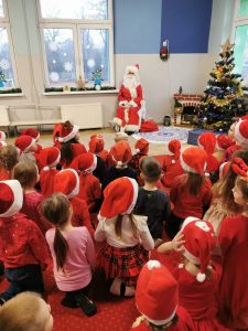 Dzieci ubrane na czerwono w mikołajkowych czapkach, siedzą na dywanie i przyglądają się świętemu Mikołajowi siedzącemu obok choinki i kominka.