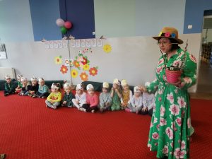 Pani Kamila w zielonej sukni w kwiaty i wiosennym kapeluszu zaprasza dzieci do obejrzenia przedstawienia. Dzieci grupy "Biedronki" siedzą w szeregu na dywanie, na głowach mają kolorowe, wiosenne opaski.