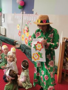 Pani Kamila (Pani Wiosna) wyciąga z koperty w kolorowe kwiaty zagadki dla dzieci. Obok znajdują się dzieci z grupy "Biedronek".