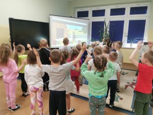 Dzieci stojÄ… w sali multimedialnej, wykonujÄ… zadania wg polecenia wyÅ›wietlanego za pomocÄ… projektora. 