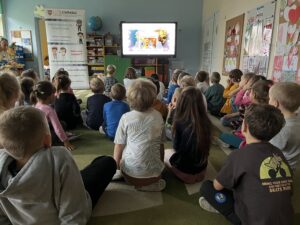 Dzieci siedzące na dywanie, oglądają film edukacyjny wyświetlany na tablicy multimedialnej.