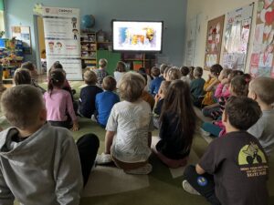 Dzieci siedzące na dywanie, oglądają film edukacyjny wyświetlany na tablicy multimedialnej.