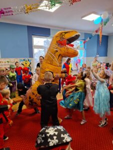 Grupa dzieci przebranych w kostiumy karnawałowe, tańcząca wokół nadmuchanego dinozaura.
