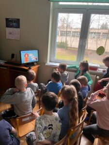 Dzieci siedząc na krzesełkach w gromadce przed monitorem komputera, oglądają film edukacyjny.