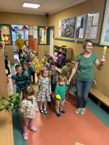 Dzieci trzymając w rękach kwiaty, podążają korytarzem przedszkola.
