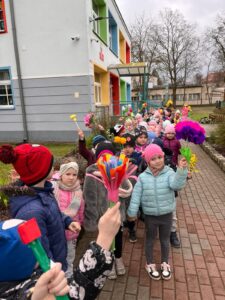 Wiosenny pochód ulicami miasta: dzieci idące w parach z kwiatami.