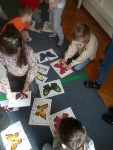 Dzieci siedząc na dywanie, układają z części w całość papierowe motyle.